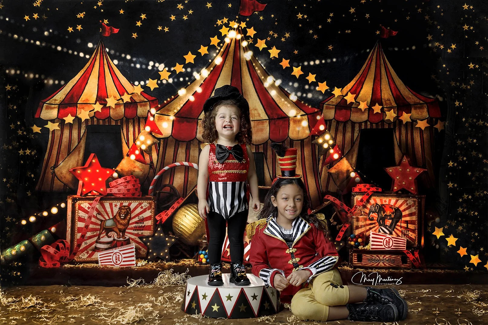 Two children dressed in circus-themed costumes, with a girl standing on a circus drum and a boy sitting on the ground, in front of a decorated circus tent backdrop with star-shaped lights and carnival props.