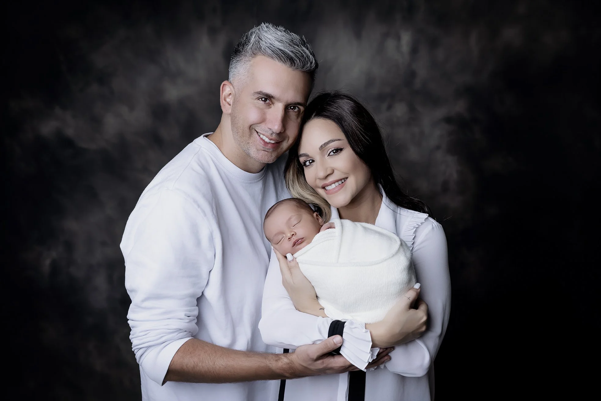 A smiling man and woman holding a sleeping baby wrapped in a white blanket, all dressed in white, against a dark background.
