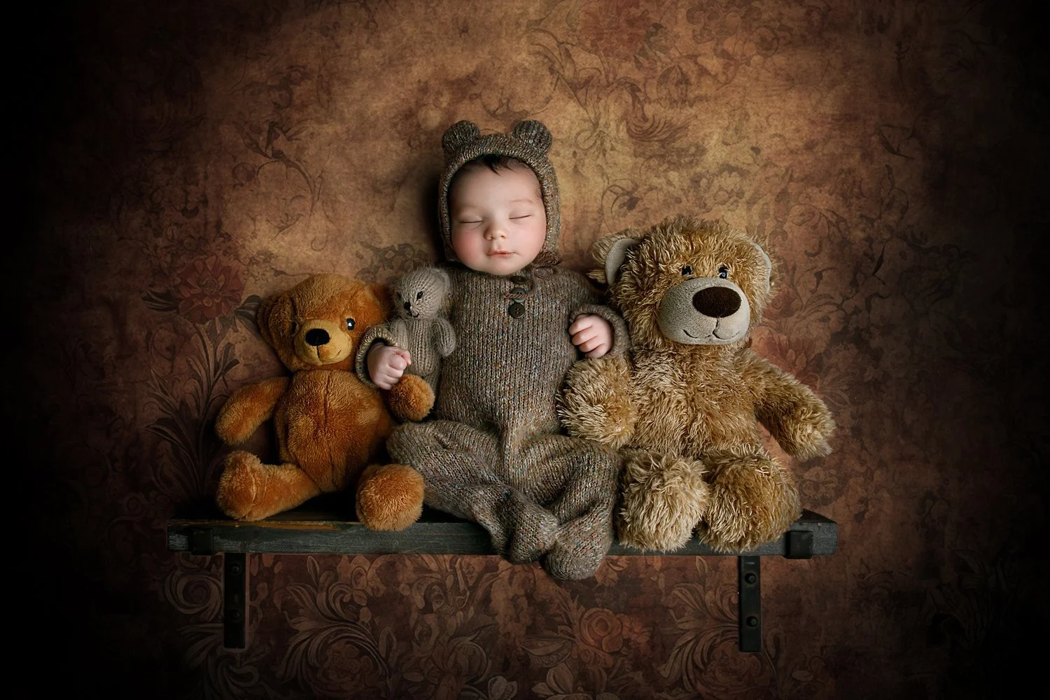 A sleeping baby dressed in a knit bear costume, surrounded by three stuffed teddy bears, sitting on a bench against a floral patterned background.