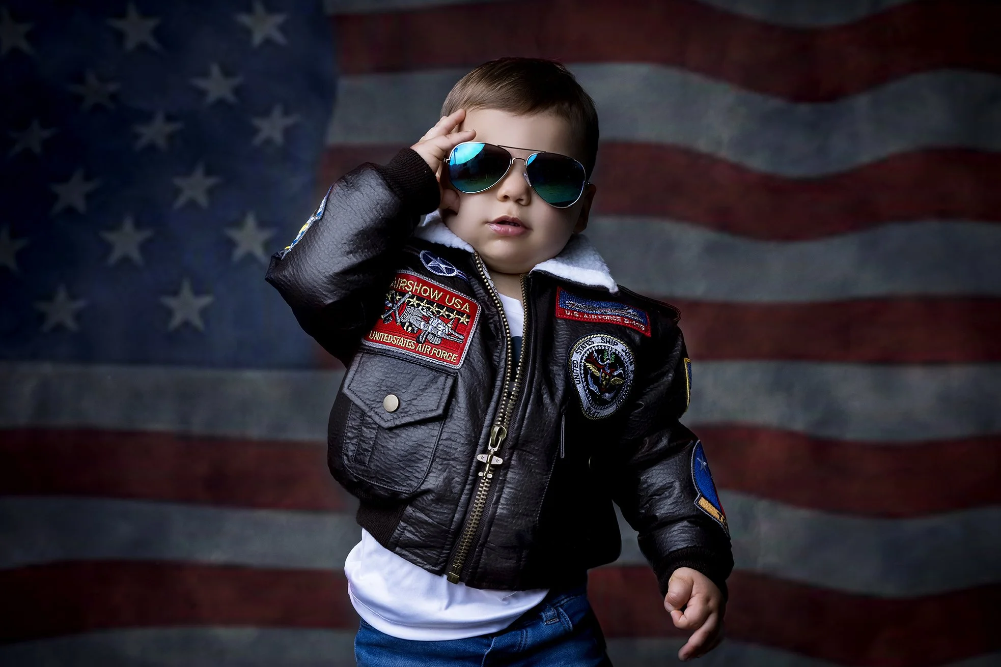 Young boy wearing aviator sunglasses, a black leather pilot jacket with patches, and a white shirt, posing in front of an American flag background.