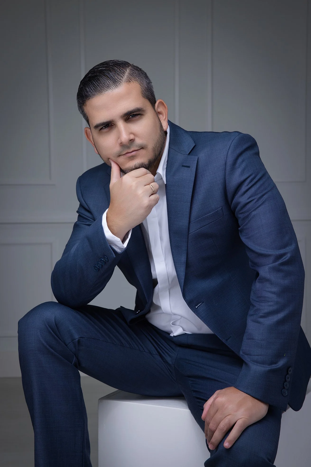 A man in a blue suit sitting on a white stool with his right elbow resting on his knee and his right hand on his chin, looking thoughtfully at the camera.