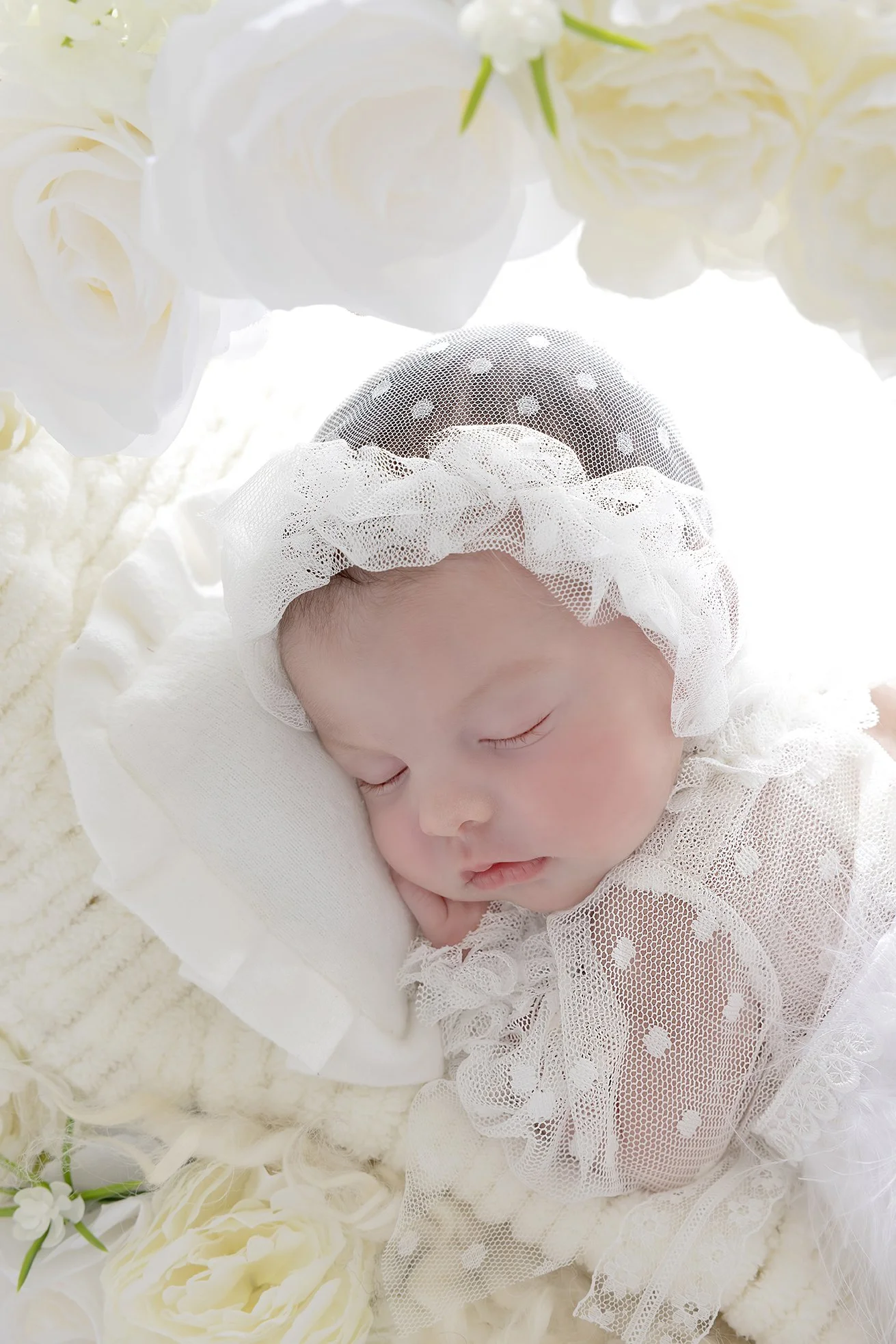 A sleeping baby wearing a white hat with polka dots and lace, resting on a white pillow, surrounded by white flowers and soft textures.