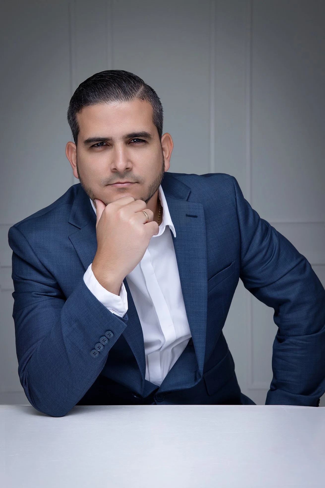 A confident man in a blue suit sitting thoughtfully at a white table against a plain wall background.