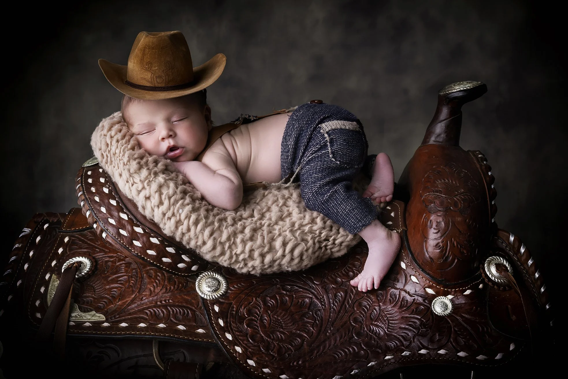 A baby sleeping on a decorative saddle with a brown cowboy hat on its head, curled on a cozy blanket.