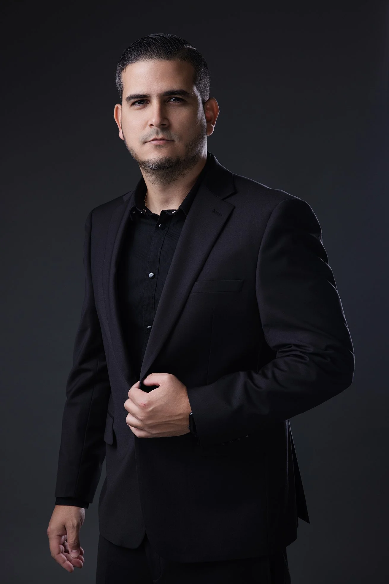 A young man in a black suit against a dark background, looking at the camera with a serious expression.