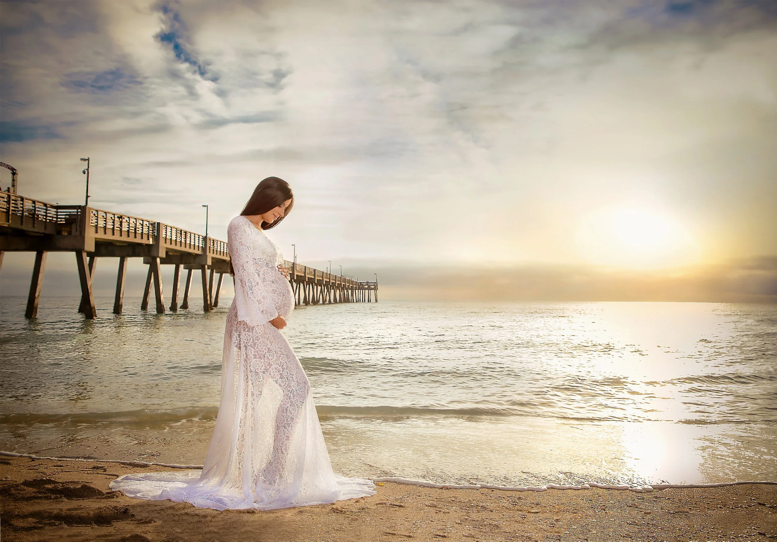 A pregnant woman in a white lace dress standing on a sandy beach at sunset, looking down at her belly with a pier in the background.