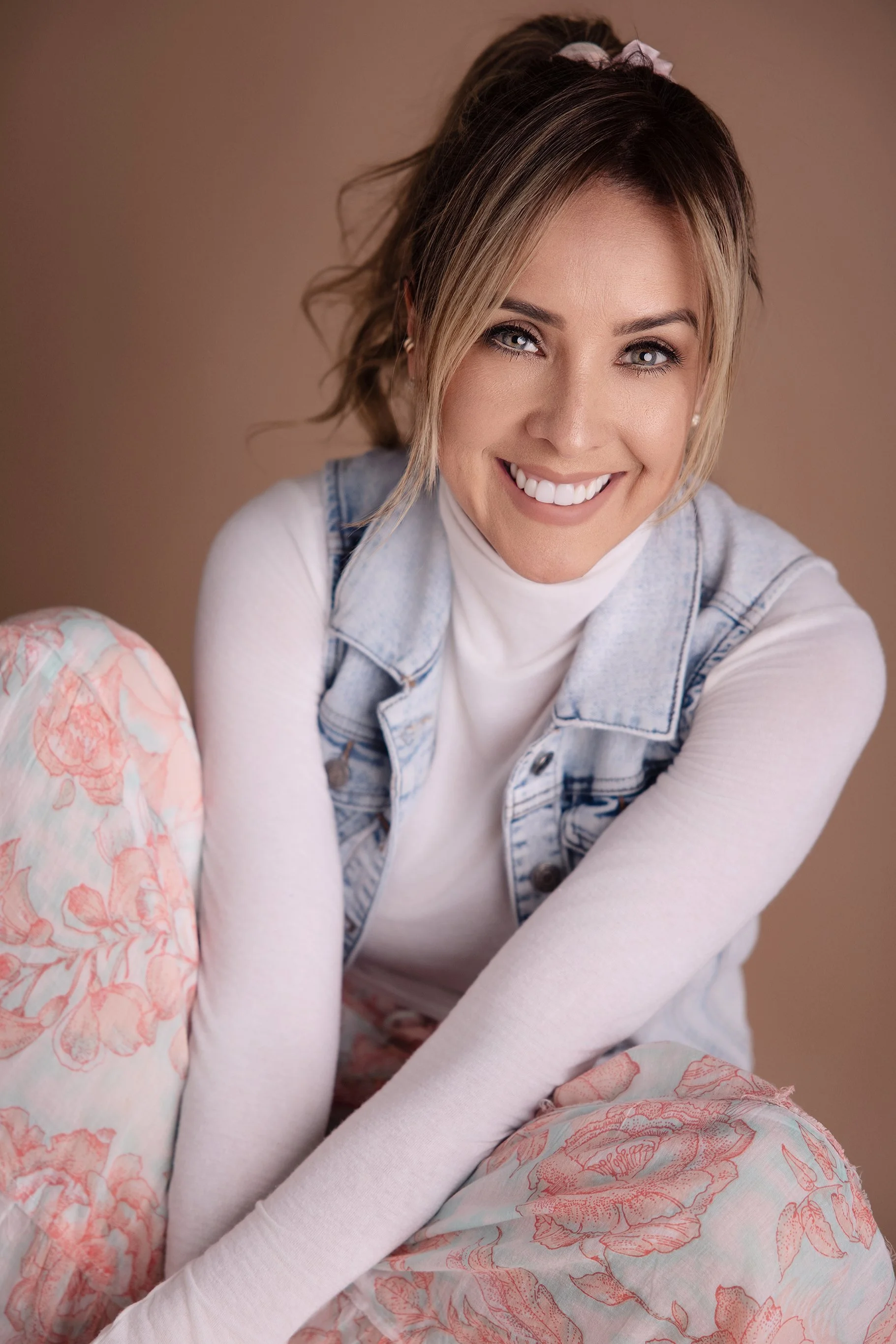 A smiling woman with blonde hair in a ponytail, wearing a white turtleneck, denim vest, and floral pants, sitting on the floor against a neutral background.