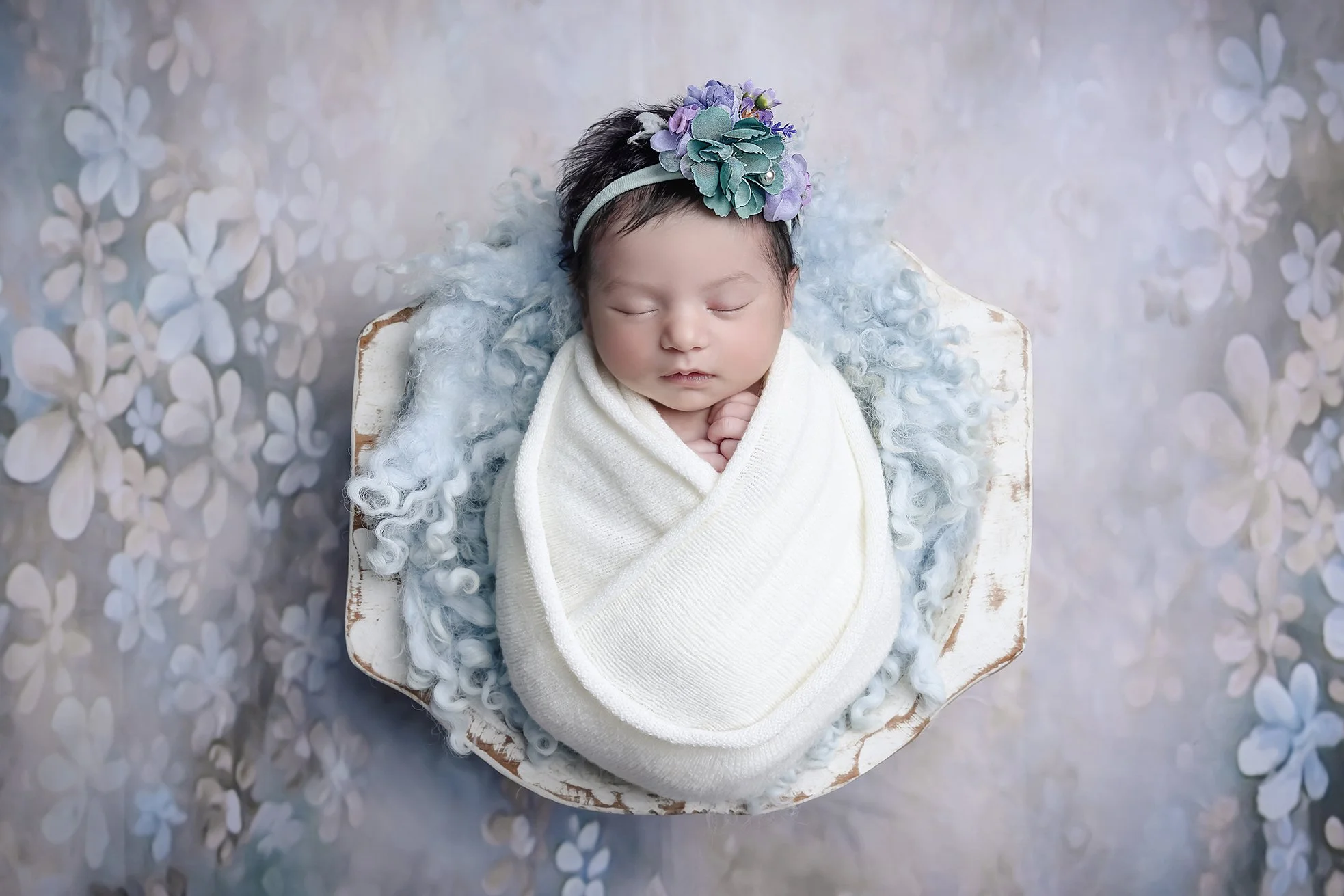 A sleeping newborn baby wrapped in a white blanket, wearing a headband with purple and teal flowers, lying on a blue curly blanket inside a white wooden basket with a floral patterned background.