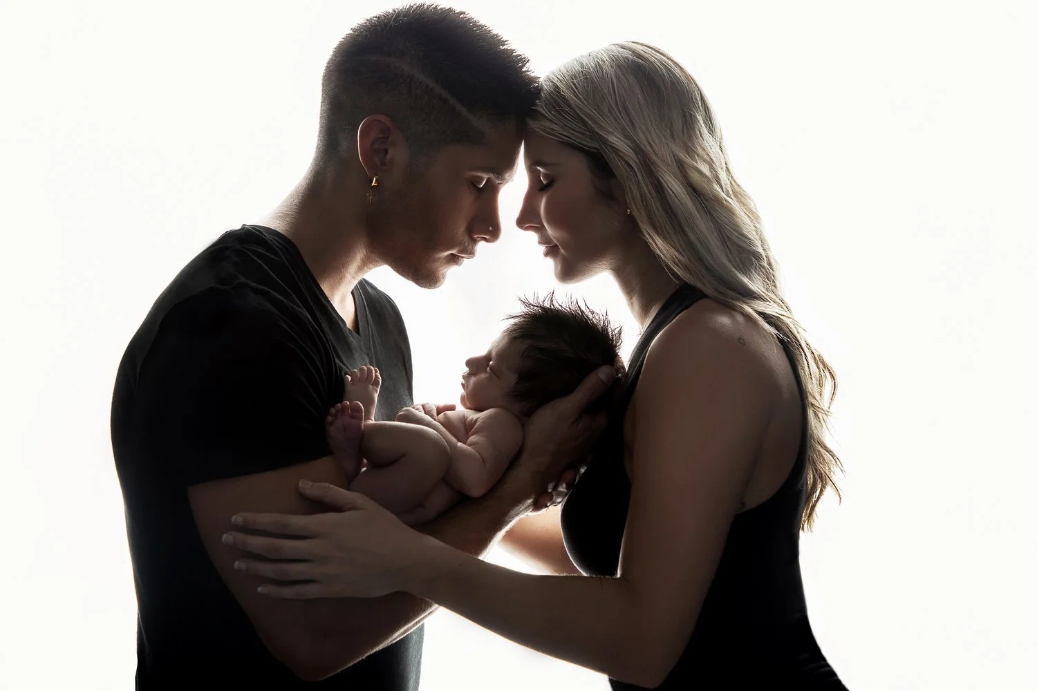 A young family of three, a man and a woman holding their newborn baby, with their foreheads touching in a tender moment, silhouetted against a white background.
