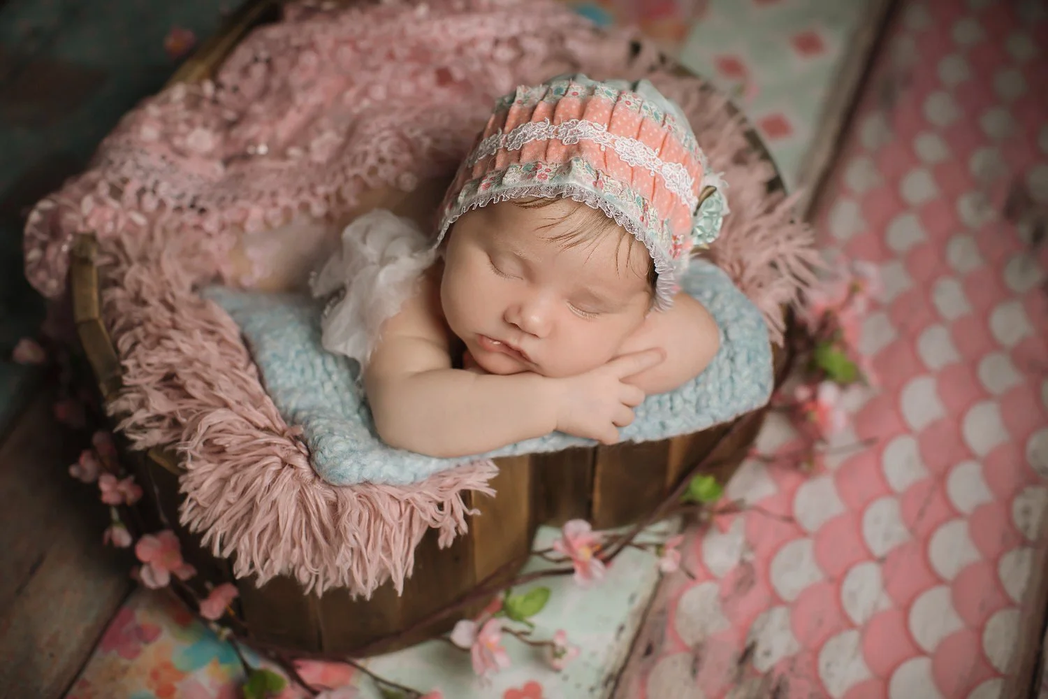 A sleeping baby wearing a pink and blue bonnet, resting on its arm inside a wooden basket, surrounded by pink flowers and fabric with heart patterns.
