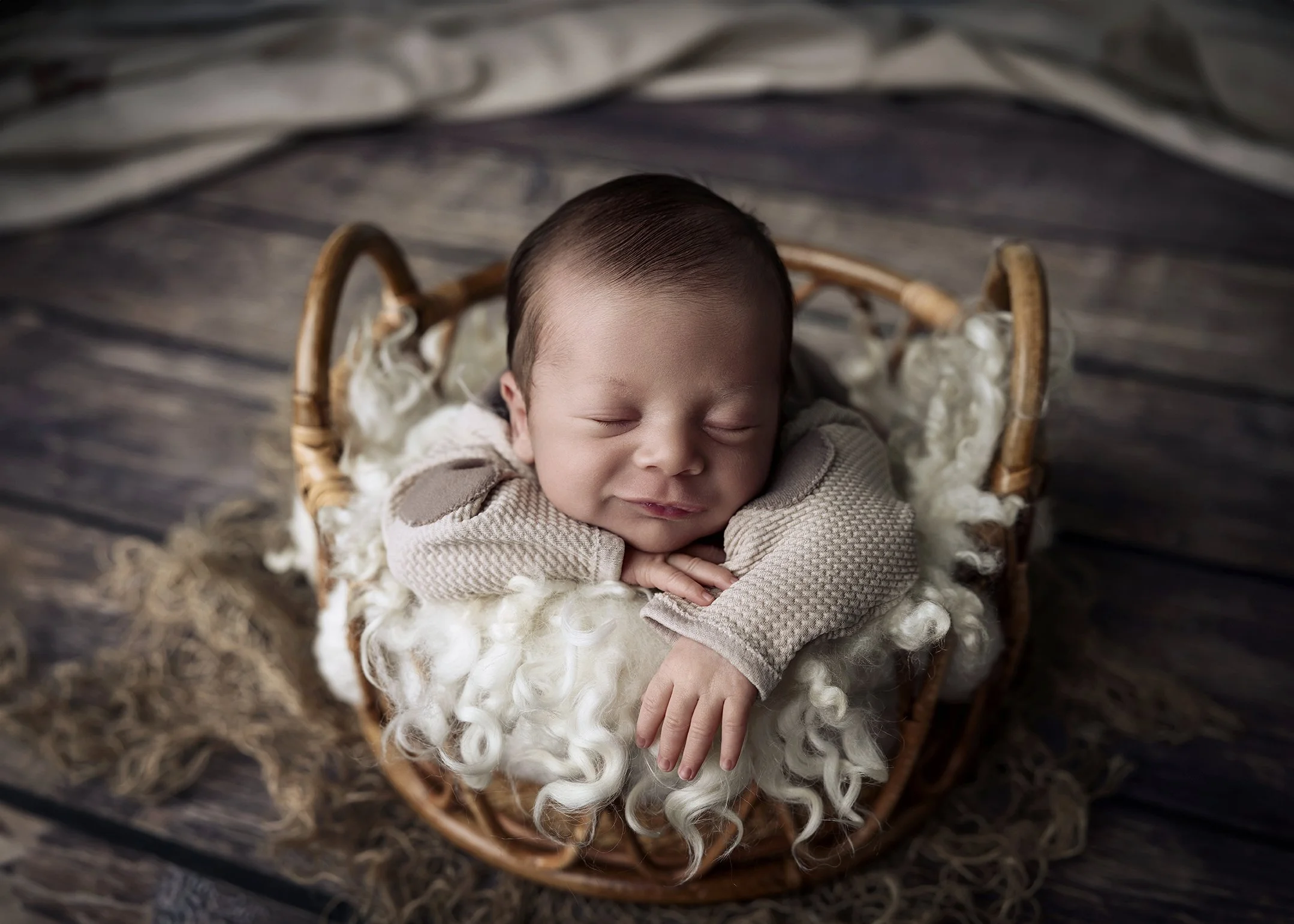 Newborn baby sleeping in a wicker basket on a wooden floor, wrapped in a soft, curly blanket.