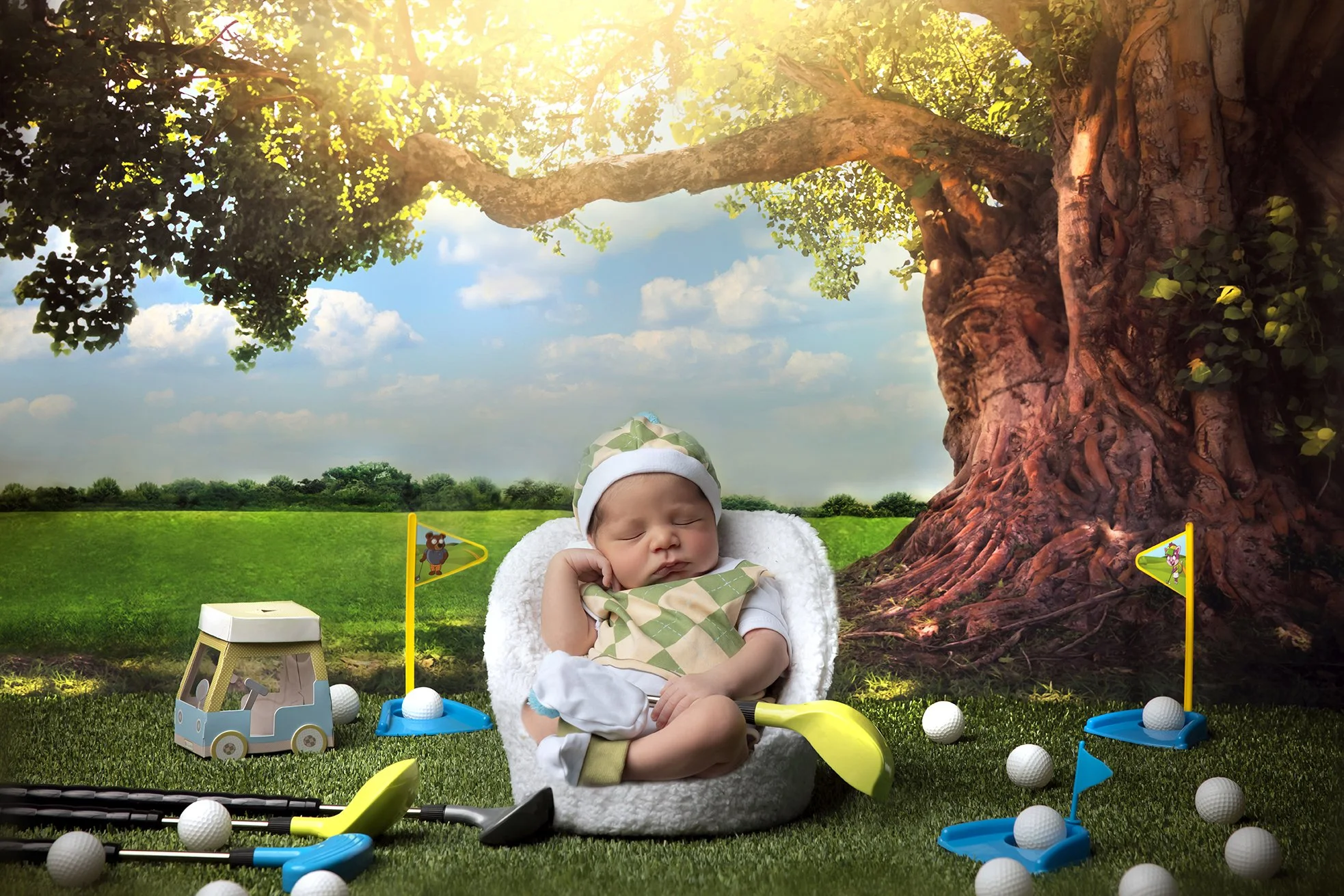 A sleeping baby with a golf-themed outfit sits in a cozy white chair outdoors under a large tree, surrounded by toy golf clubs, golf balls, and flags in a vibrant green field on a sunny day.