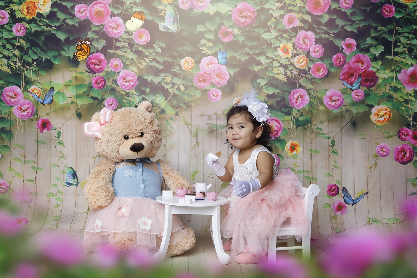 A young girl in a pink tutu dress and white floral headband sits at a small white table, drinking from a teacup, with a large teddy bear dressed in a pink flower dress and bow sitting nearby. The background features pink flowers and butterflies on a 