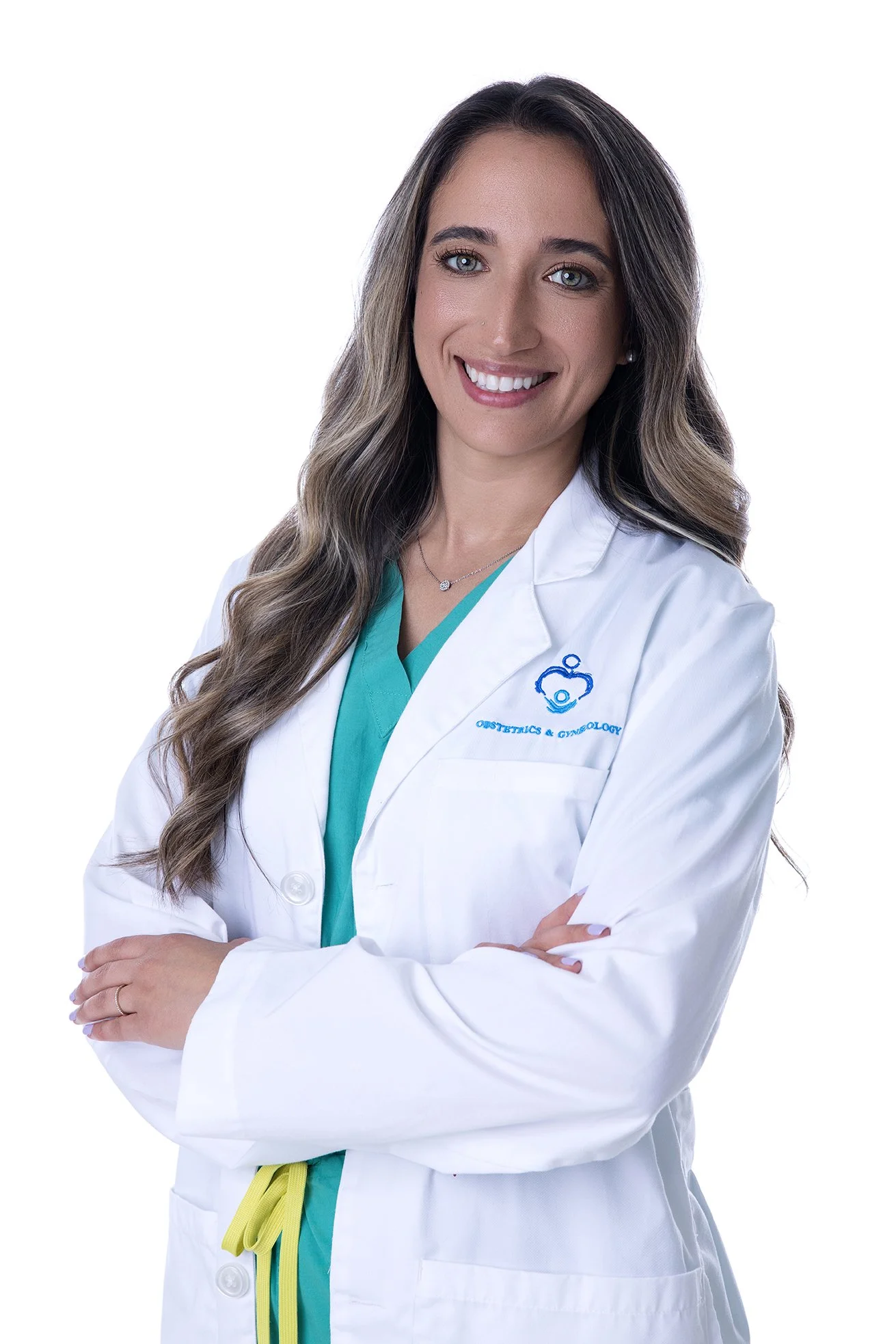 A female doctor in a white coat with her arms crossed, smiling, with long wavy hair, wearing a teal scrubs underneath, and standing against a white background.