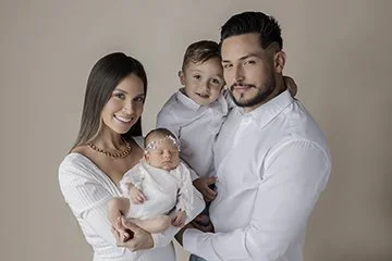 Family portrait of a man, woman, and two children, including a baby, against a plain background.