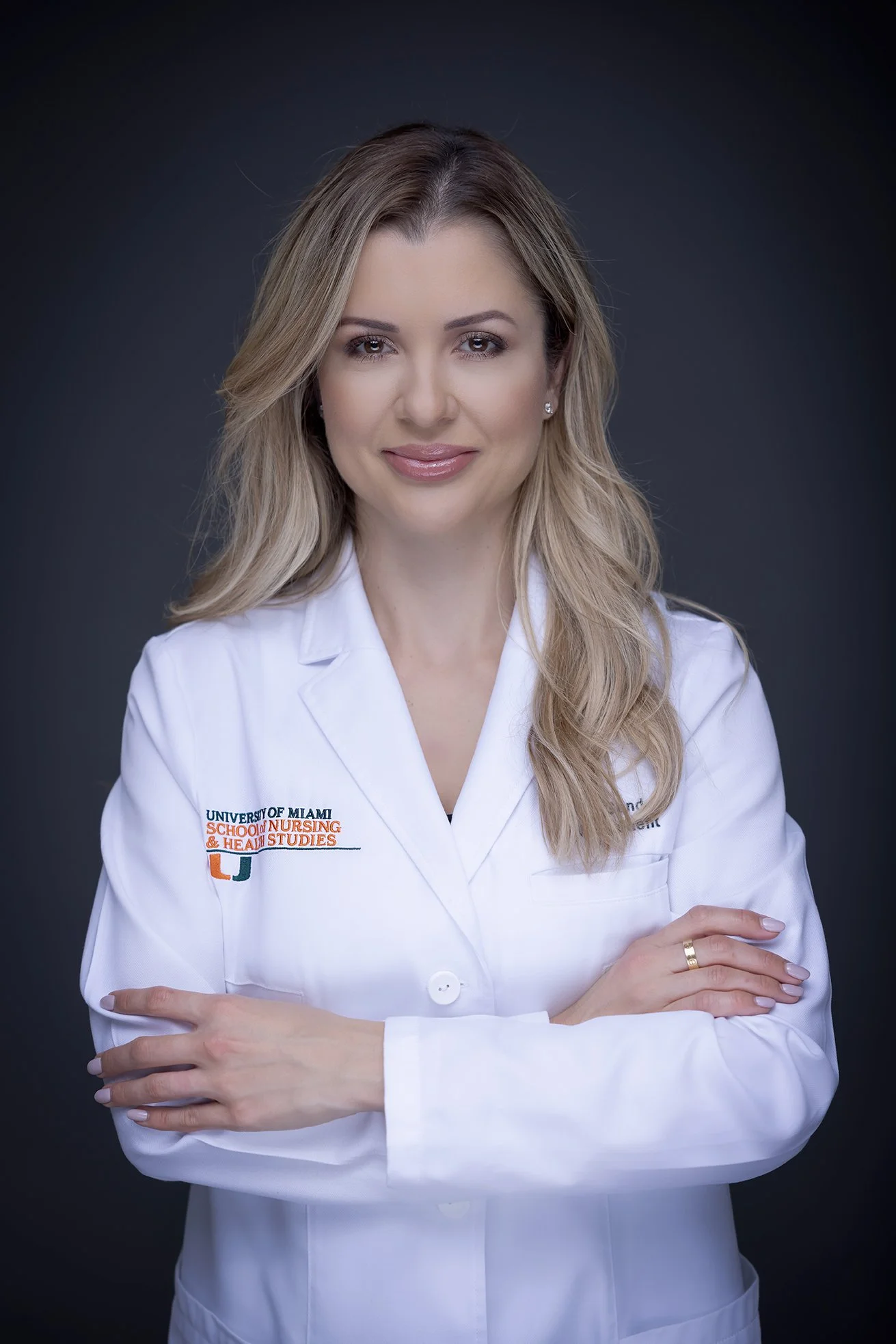 A woman in a white medical coat with 'University of Miami School of Nursing & Health Studies' logo standing against a dark background.