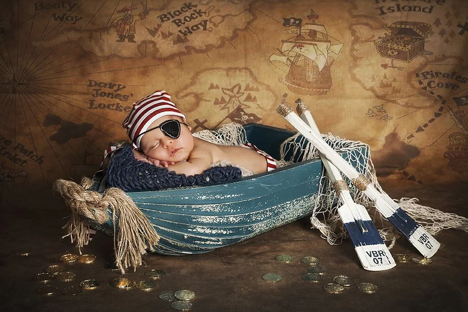 A baby dressed as a pirate, wearing a striped pirate hat and an eye patch, is sleeping in a small blue boat with a backdrop of a pirate-themed map. The boat is filled with blankets, and there are pirate swords and coins around it.