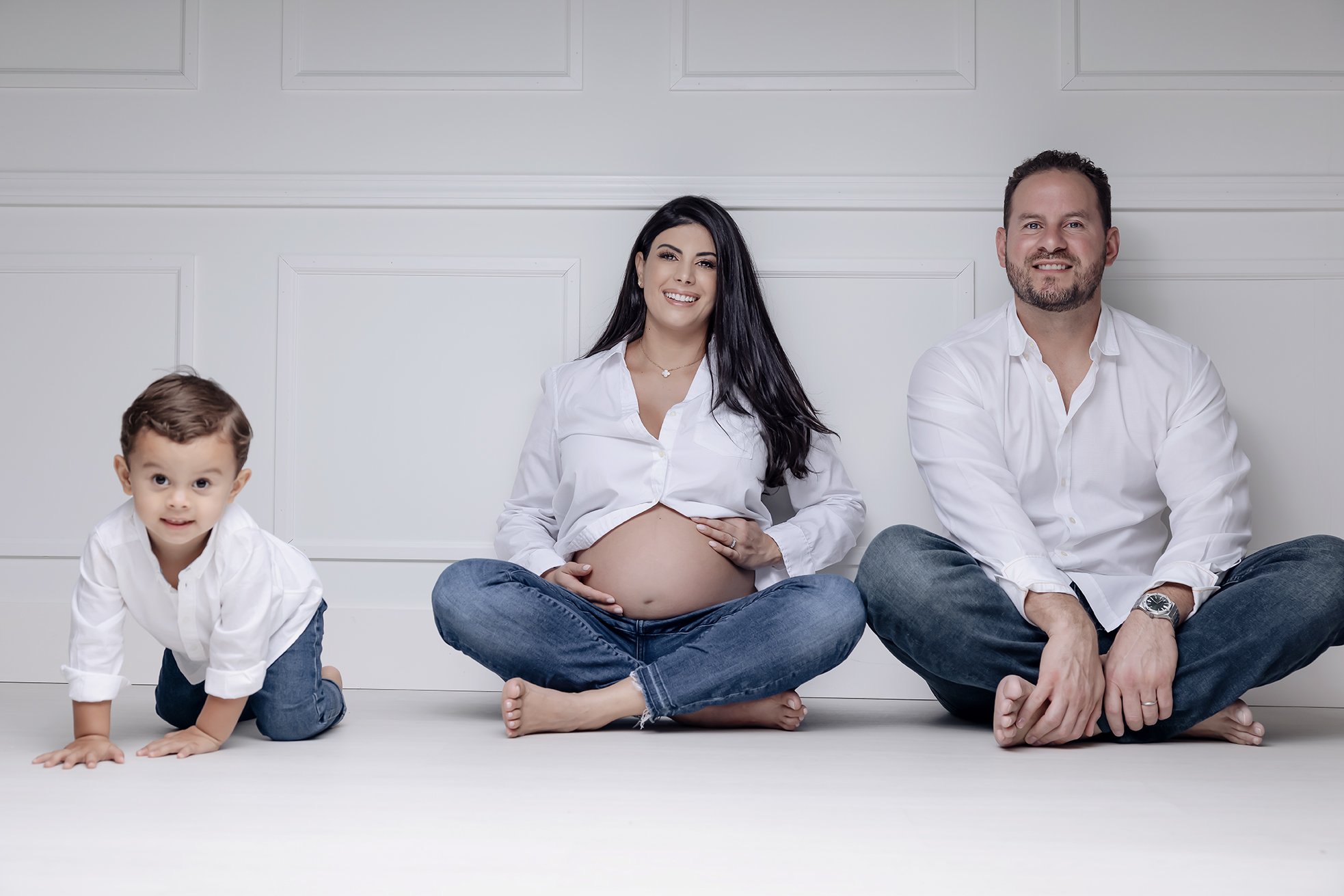 A pregnant woman, a man, and a young boy sitting on the floor against a white wall, all dressed in white shirts and jeans.