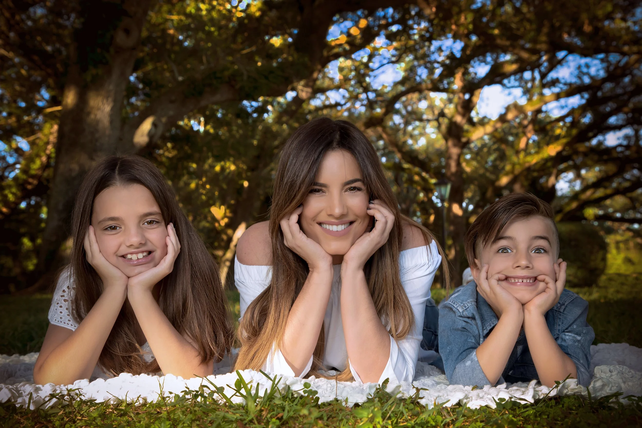 A woman and two children lying on the grass outdoors during daytime, smiling and resting their faces on their hands, with trees and sunlight in the background.