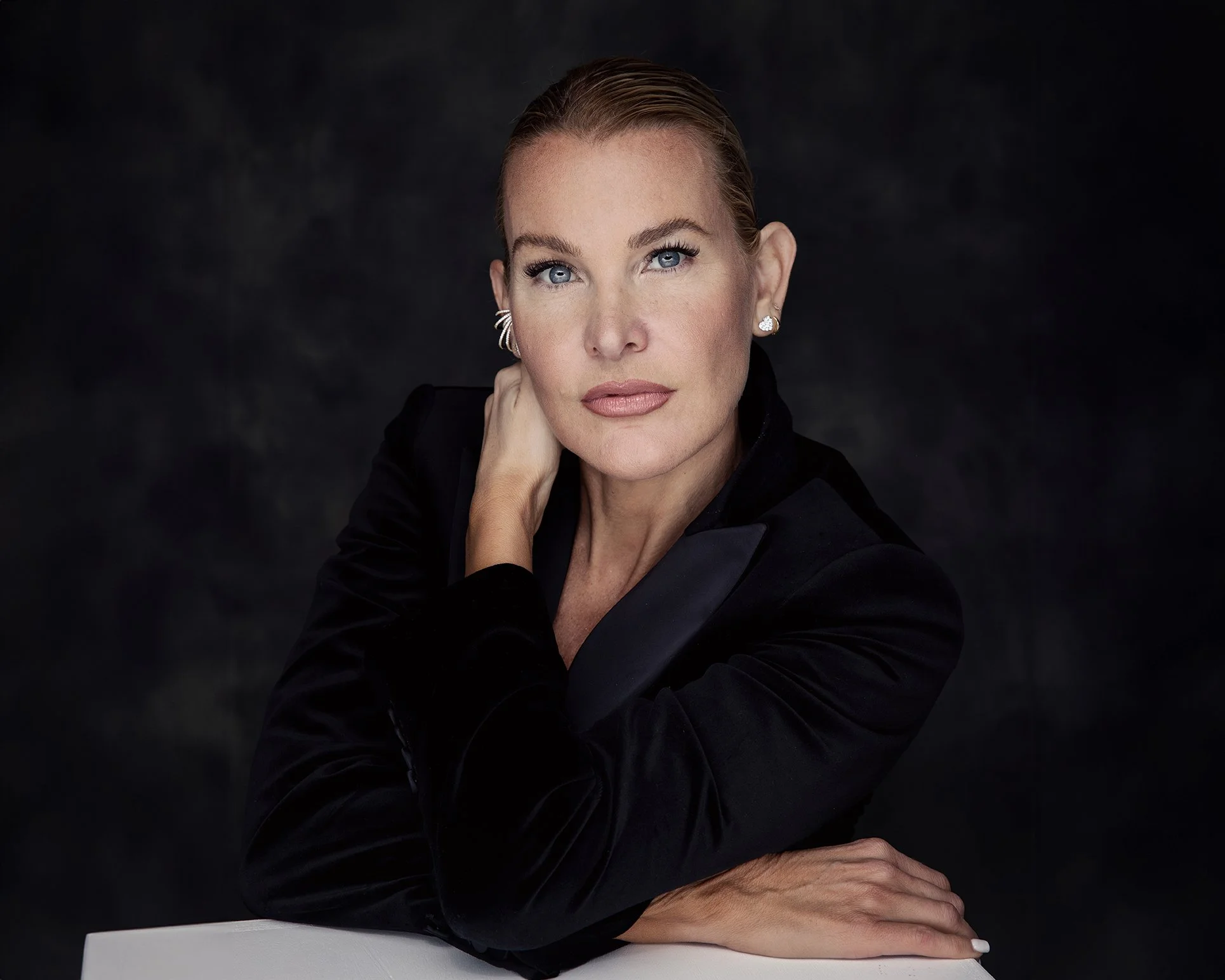 A woman with blonde hair, blue eyes, and wearing a black blazer, resting her chin on her hand against a dark background.