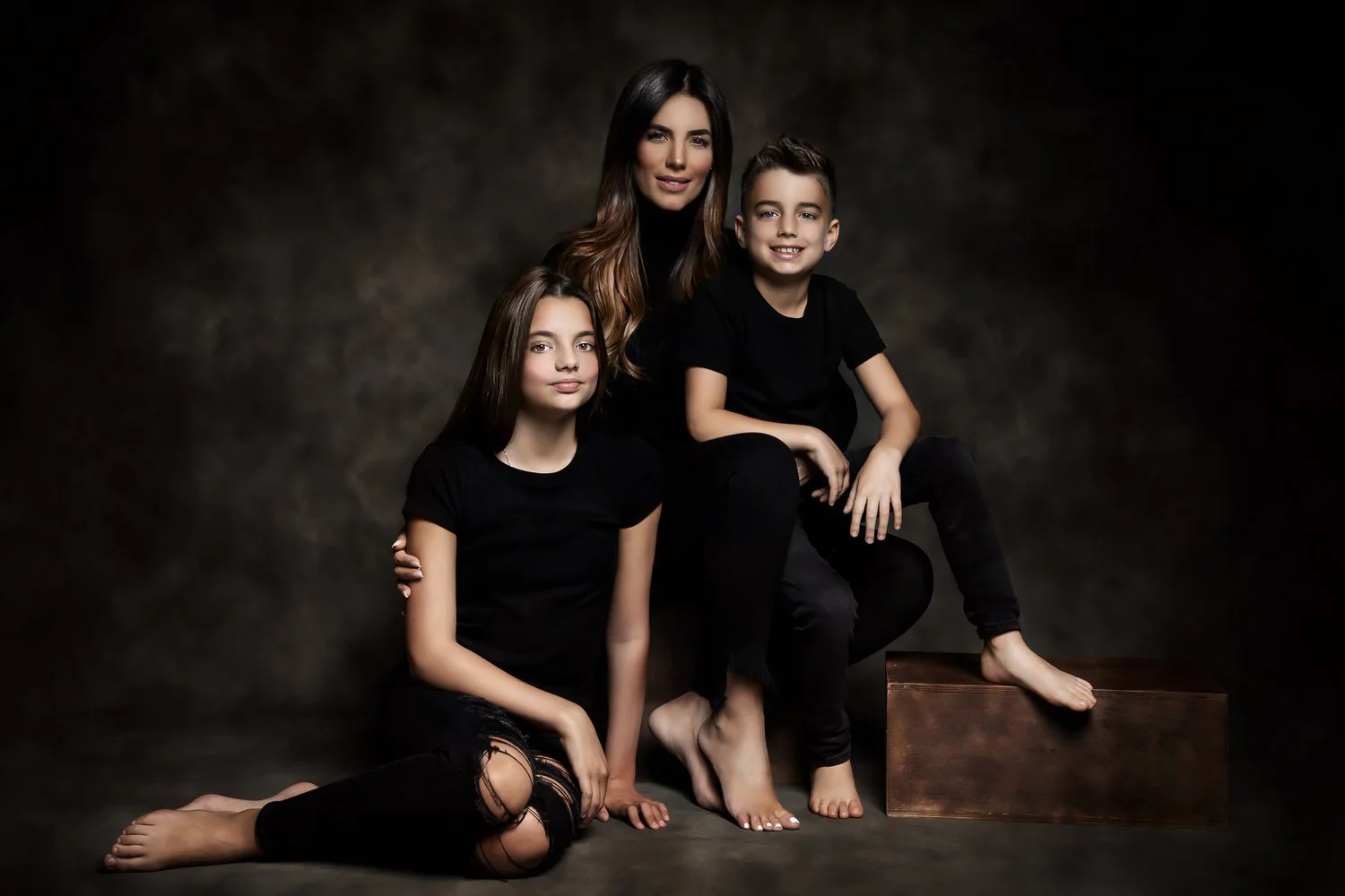 A woman and two children in black clothing posing for a studio portrait against a dark backdrop. The woman has long brown hair, and the girl has long dark hair and is sitting on the floor with her legs crossed. The boy has short dark hair, is sitting