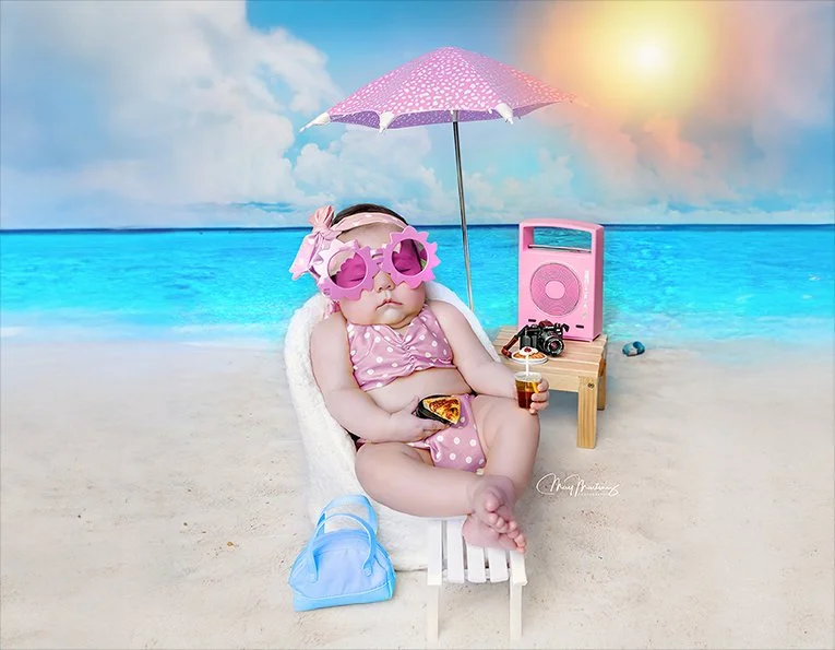 Child in pink polka dot swimsuit and large pink sunglasses sitting in a beach chair under a pink umbrella, with a mini radio, camera, and drink nearby, on a sandy beach with ocean and sky in the background.