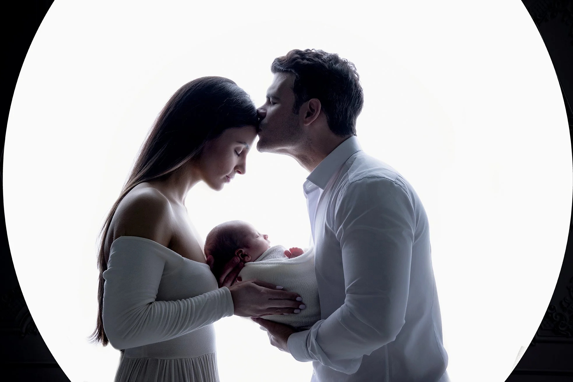 A family of three with a mother, father, and newborn baby, with the father kissing the mother's forehead while she holds the baby.