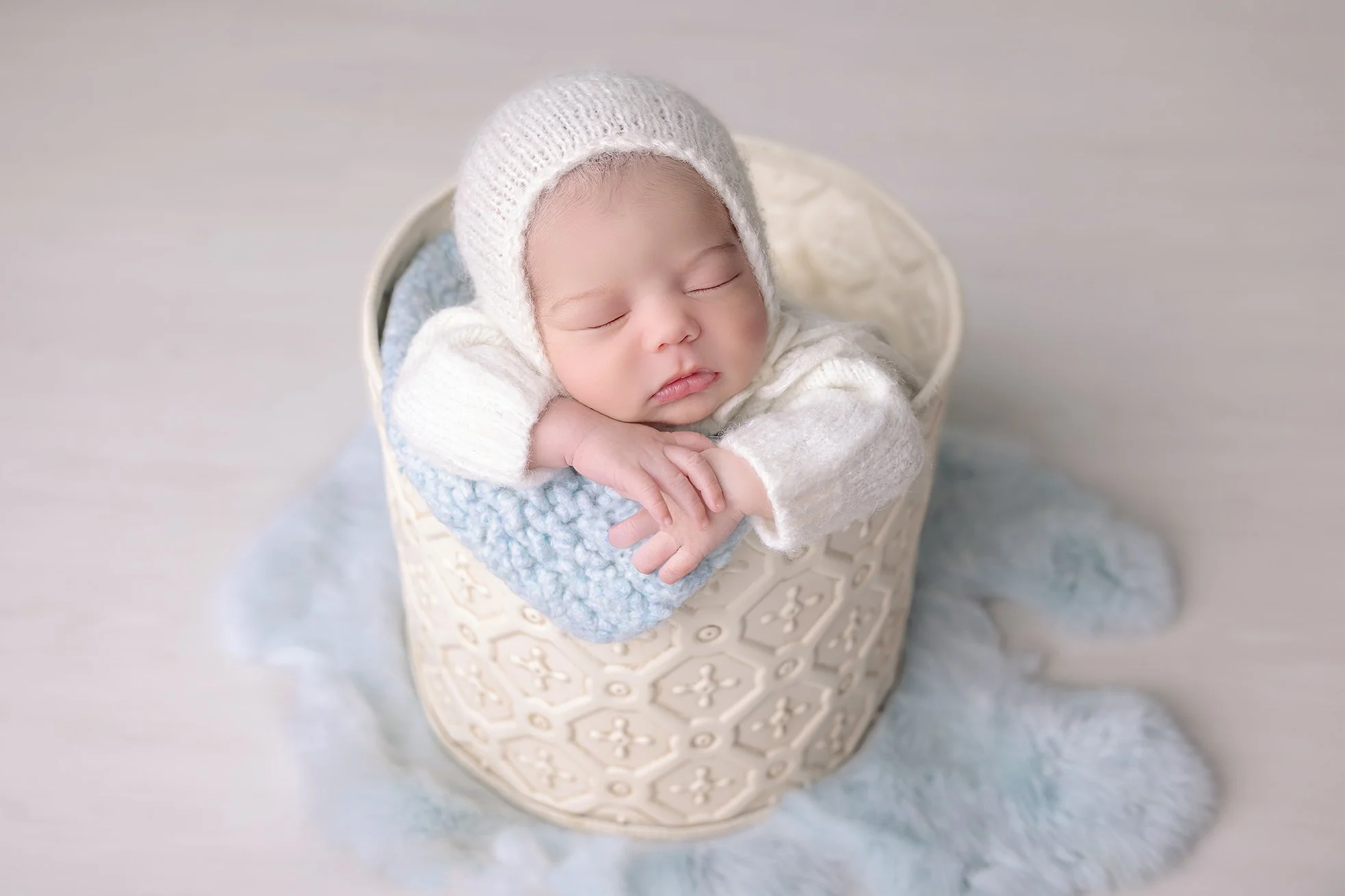 A sleeping baby wearing a knit hat and sweater, resting their head on their arms inside a basket on a soft, fluffy, blue blanket.