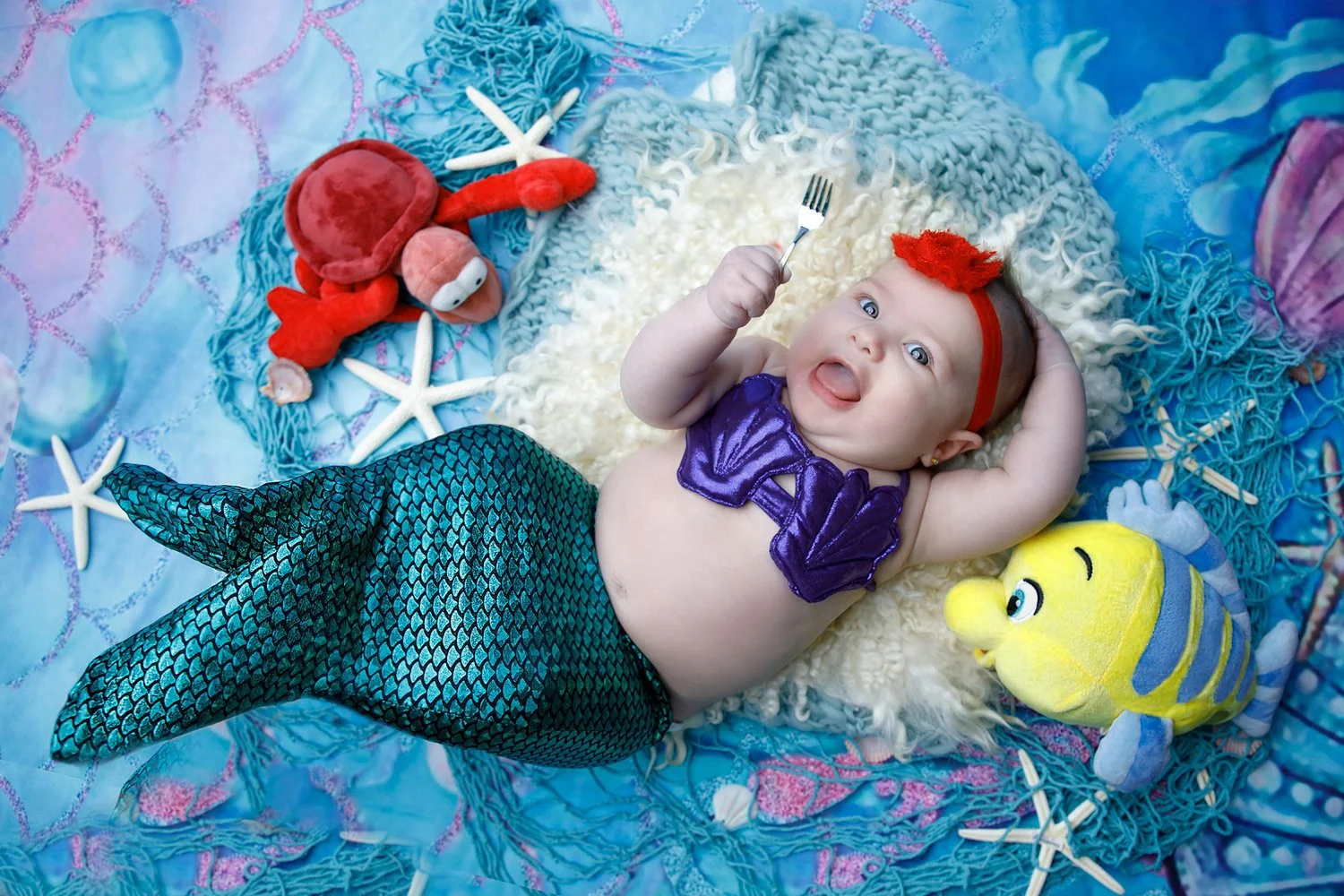 Baby dressed as a mermaid with a purple top and a shiny blue-green tail, lying on an ocean-themed blanket surrounded by plush toys of sea creatures including a red crab, a yellow and blue fish, and white starfish, holding a small fork in one hand.
