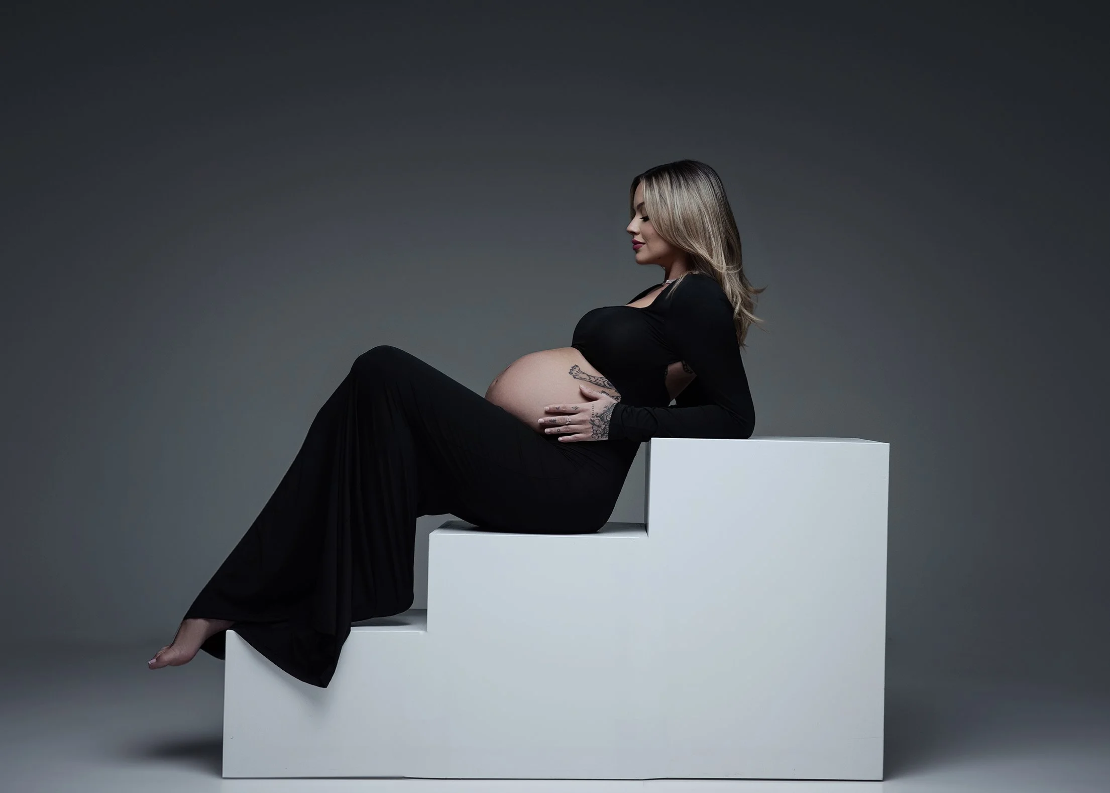 Pregnant woman with tattoos sitting on a white block against a gray background, wearing a black dress and holding her belly.