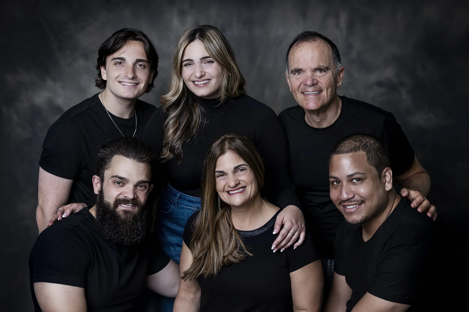 Group portrait of seven people, three women and four men, posing together against a dark, textured background, all wearing black tops and smiling.