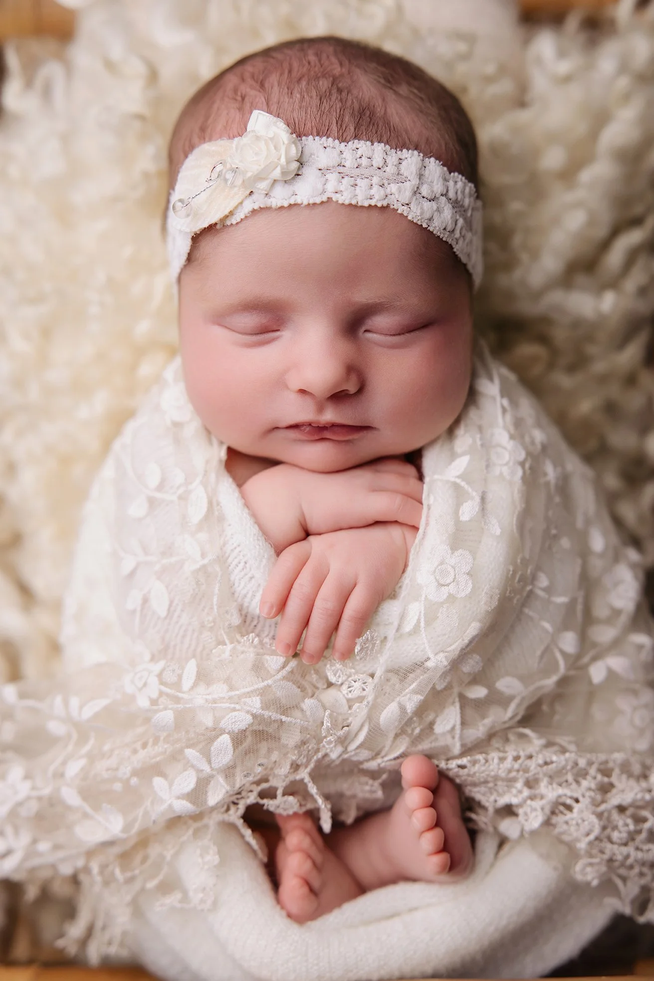 A sleeping baby wrapped in a lace, embroidered blanket with a floral pattern, wearing a white headband with a small flower, and lying on a soft, cream-colored surface.