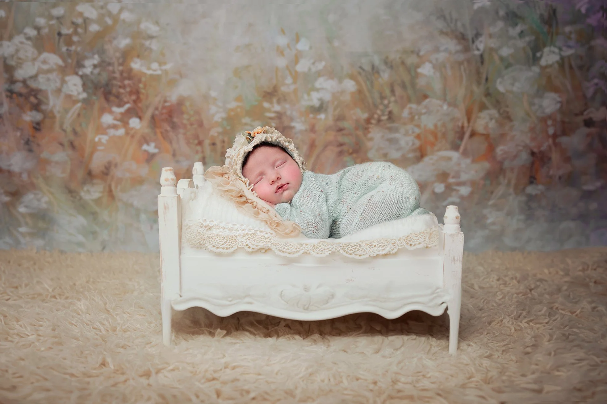 A sleeping baby lying on a small vintage-style white bed with lace trim, wearing a knitted outfit and a bonnet, in front of a colorful floral background and a beige shaggy rug.