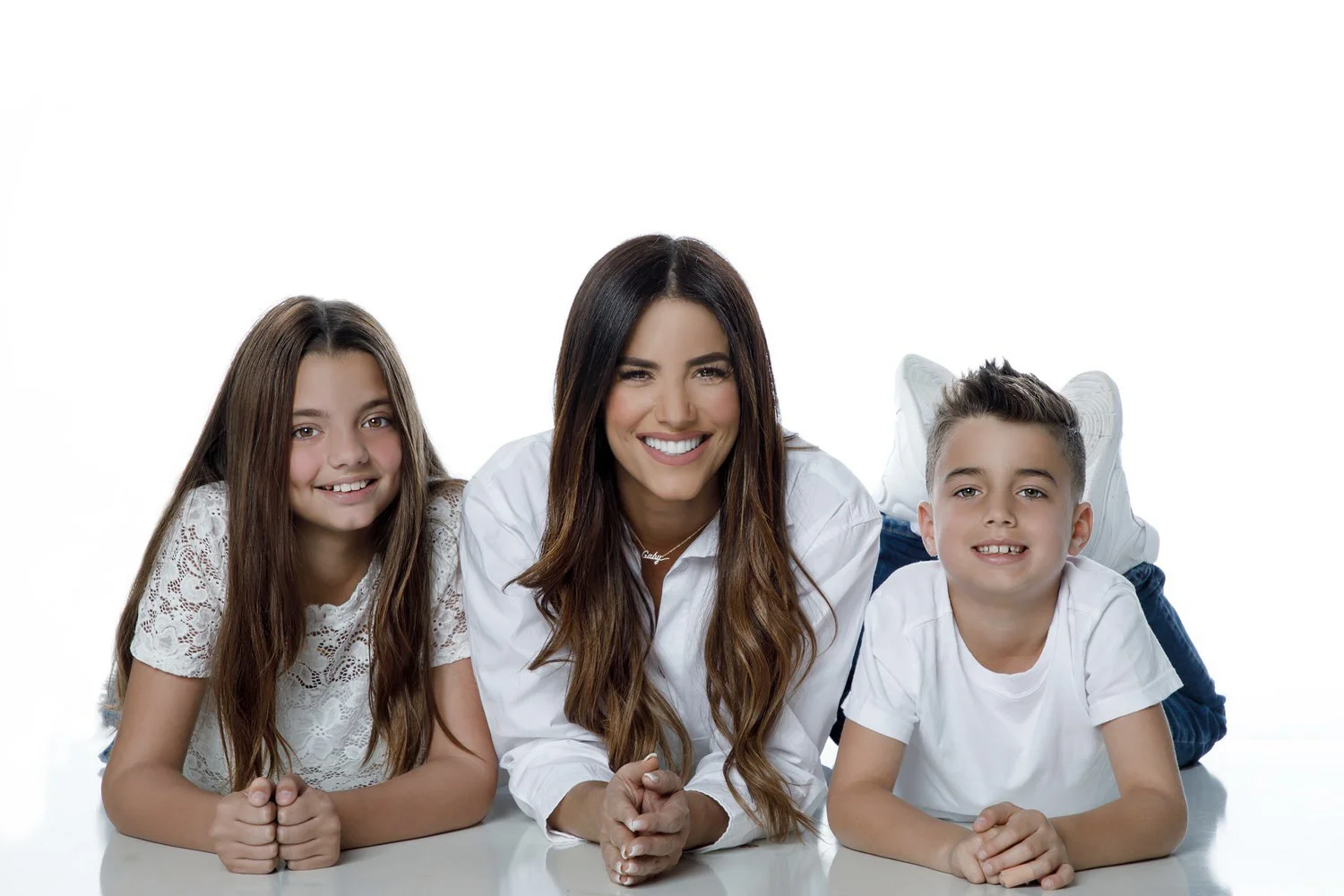 A woman and two children lying on the floor with their heads resting on their hands, smiling at the camera against a white background.