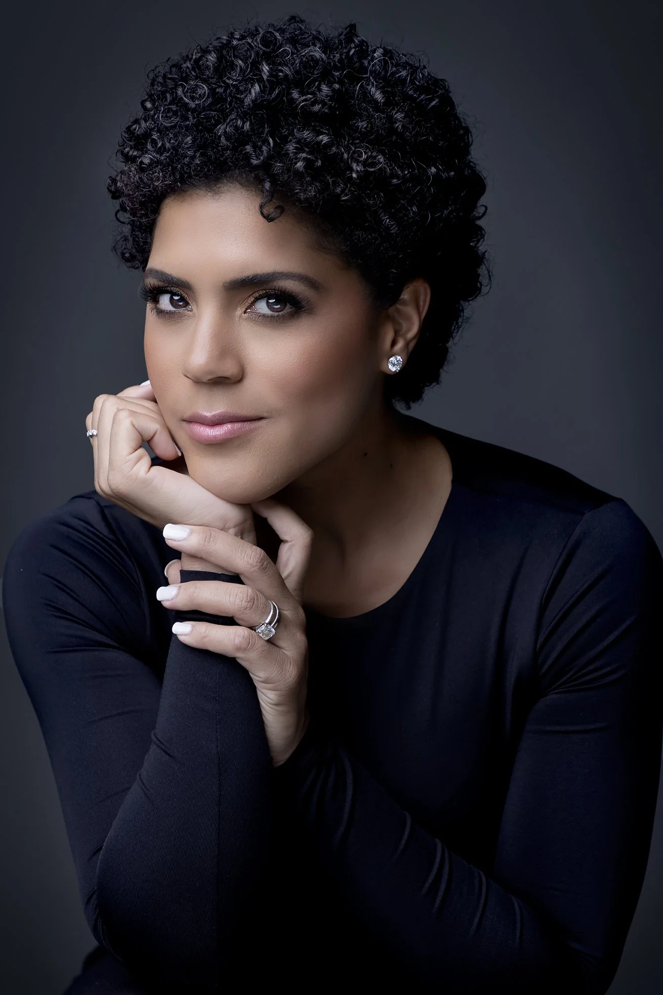 A woman with short, curly black hair and light skin, wearing diamond earrings and rings, poses with her chin resting on her hand against a dark background.