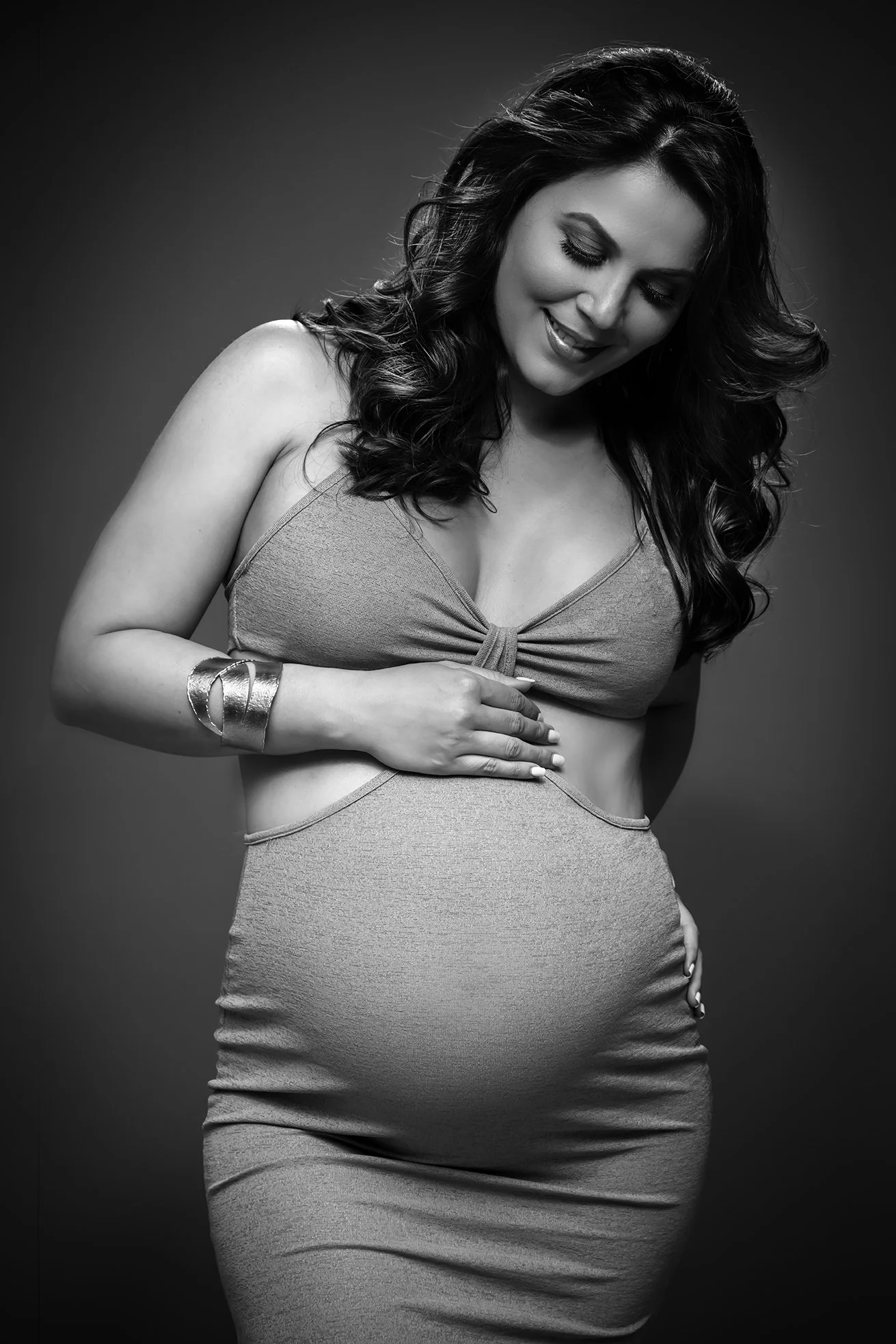 Black and white photo of a pregnant woman with wavy hair, smiling with her eyes closed, wearing a sleeveless top and a fitted skirt, holding her belly with her right hand