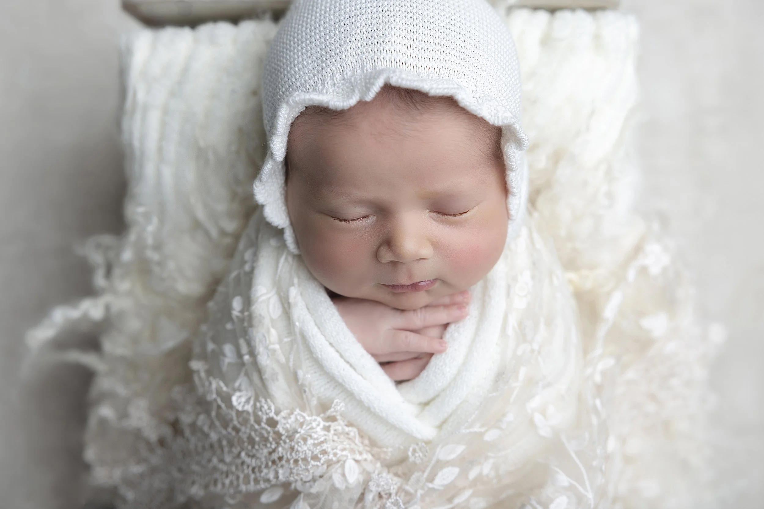 A sleeping baby wrapped in a white lace blanket, wearing a white knit hat, lying on a soft, textured white surface.