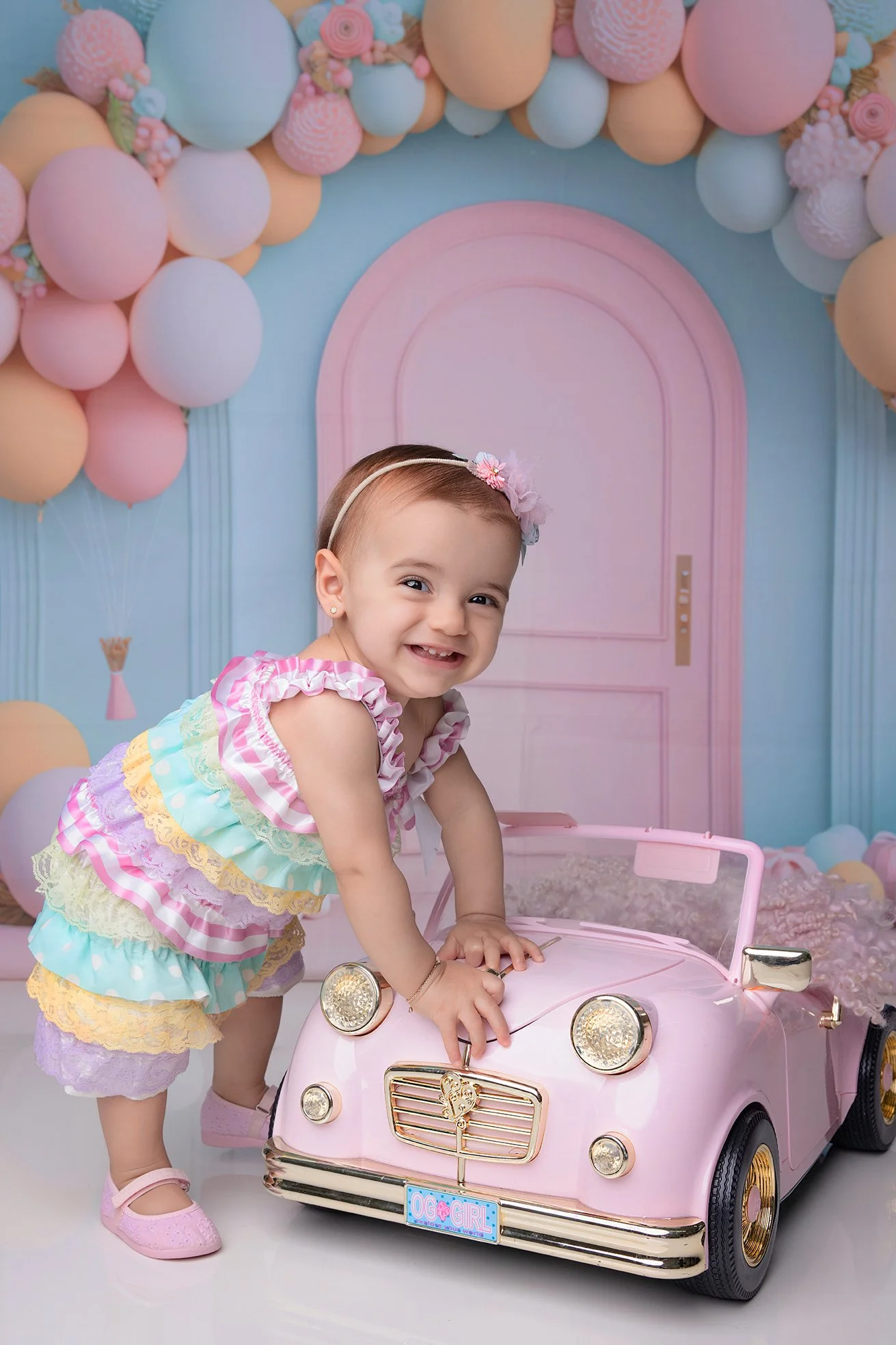 A young girl with a headband is smiling and playing with a pink toy car at a birthday celebration with pastel balloon decorations in the background.