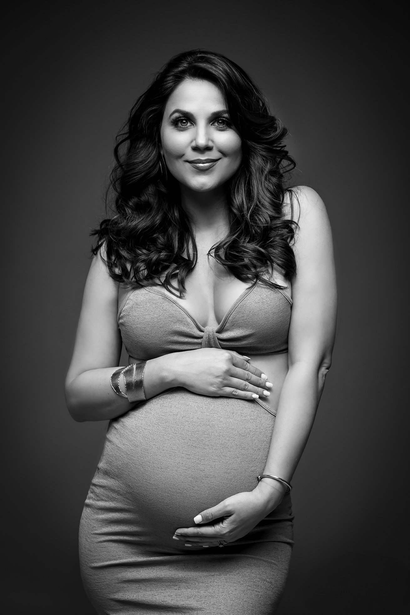 Black and white portrait of a smiling pregnant woman with long curly hair wearing a sleeveless top and a skirt, posing with hands on her belly.