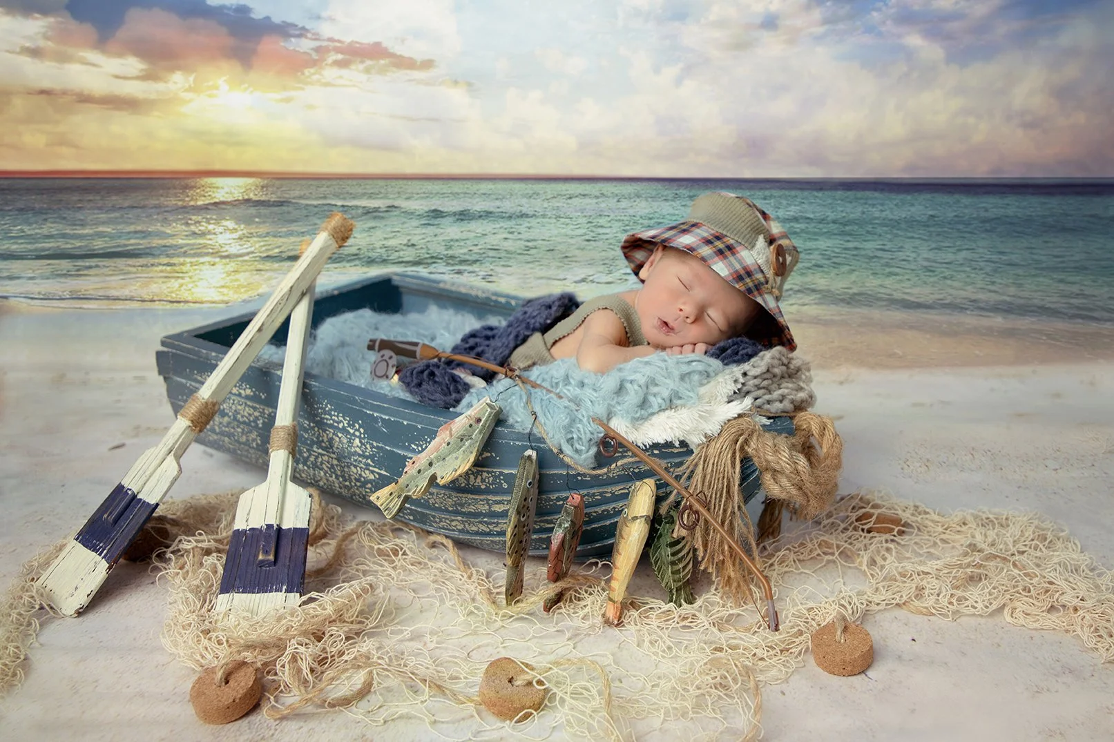 A sleeping baby in a small boat on a beach with a sunset in the background. The baby is wearing a plaid hat with a button and is surrounded by ocean-themed decorations like toy fish, oars, and fishing nets.