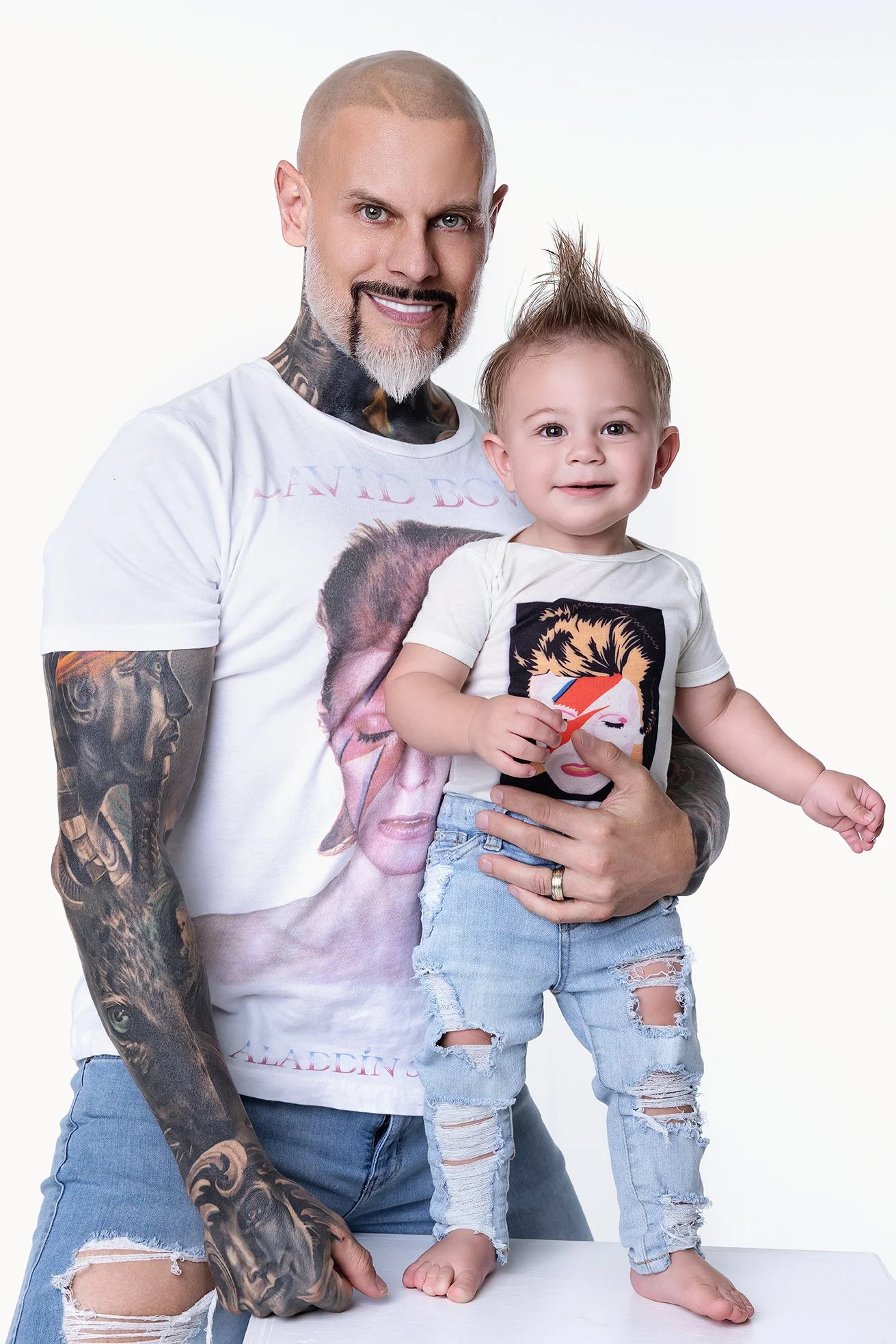 A man with tattoos holding a smiling young child with a mohawk hairstyle, both facing the camera against a plain white background.