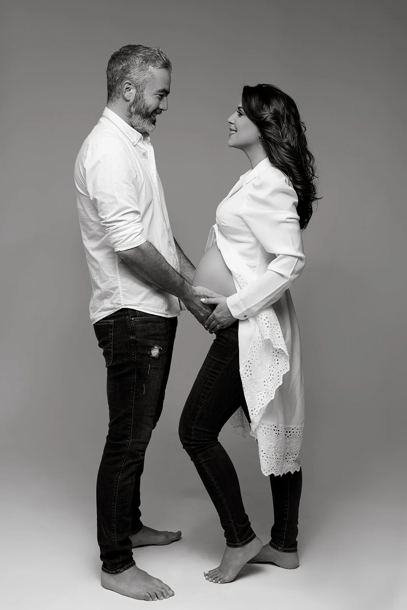 A happy couple standing close together, with the woman pregnant, holding hands and smiling at each other in a studio with a plain background.
