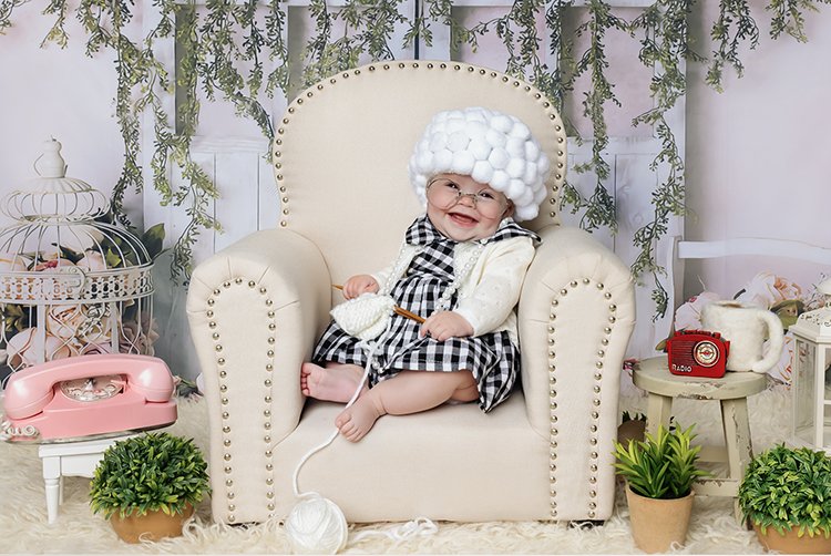 Smiling baby sitting on a cream armchair wearing a black and white checkered dress, white cardigan, and a large white fluffy hat, with a vintage telephone in hand, decorated with plants and retro items around.