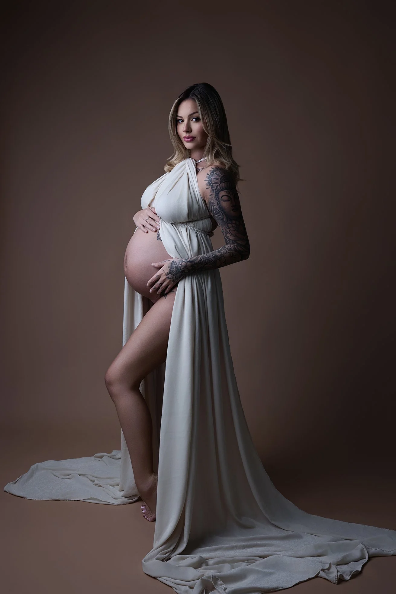 Pregnant woman with tattoos wearing a flowing beige dress, standing against a neutral background.