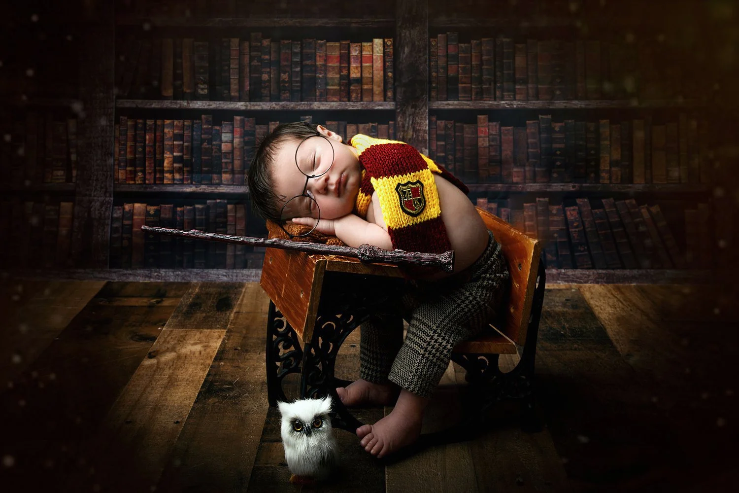 A young boy dressed as a Harry Potter character, wearing glasses and a Gryffindor scarf, sleeping on a small wooden desk. He is holding a wand while a white owl plush toy is hanging from his foot. The background resembles a library with shelves fille
