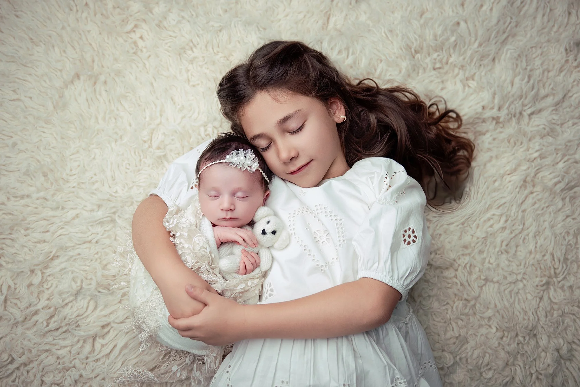A young girl and a newborn baby are lying on a cream-colored fluffy blanket, sleeping peacefully with their heads close together. The girl has long brown hair and is wearing a white dress, while the baby is dressed in a white lace outfit and a decora