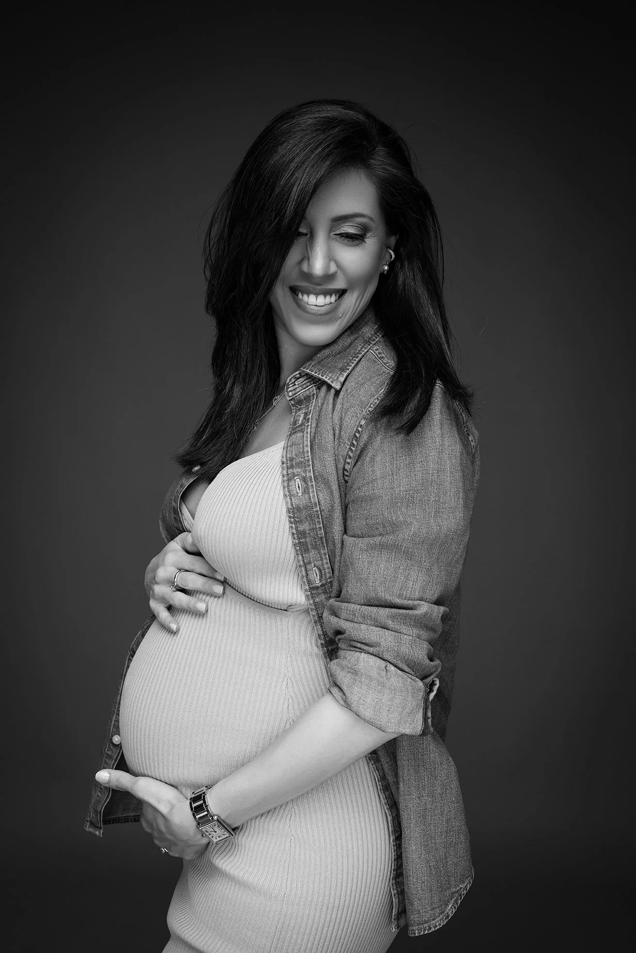 Black and white photo of a pregnant woman smiling, wearing a denim jacket over a ribbed dress, with one hand on her belly and the other holding her wrist.