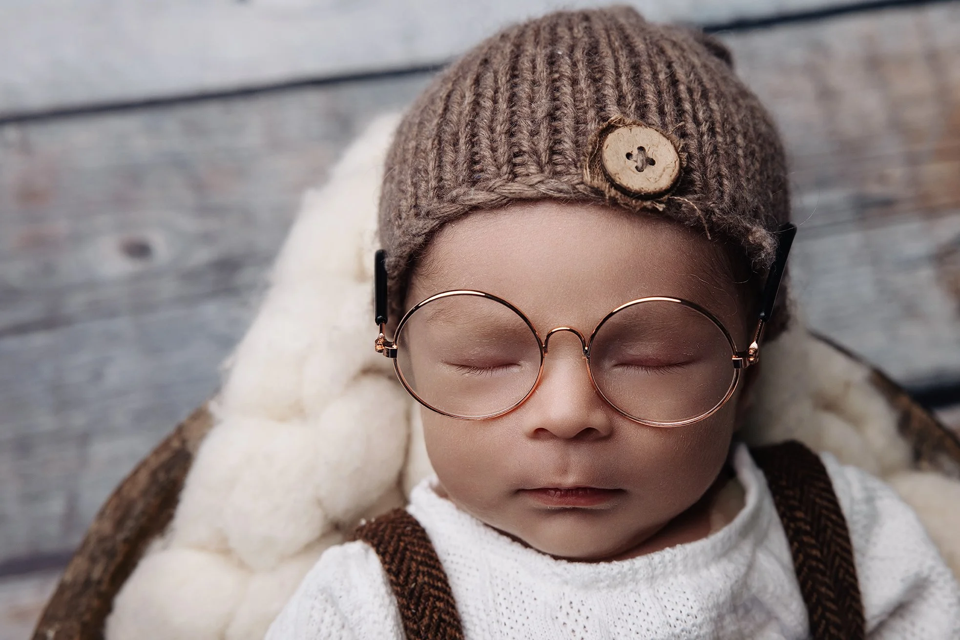 A sleeping child wearing glasses, a knitted cap with a button, and a white sweater, lying on a soft blanket on a wooden surface.