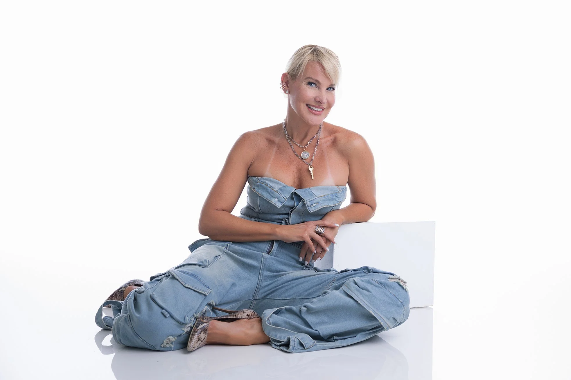 A woman with short blonde hair, wearing a strapless denim top and ripped jeans, is sitting on the floor next to a white box, smiling at the camera, with jewelry including necklaces and earrings.