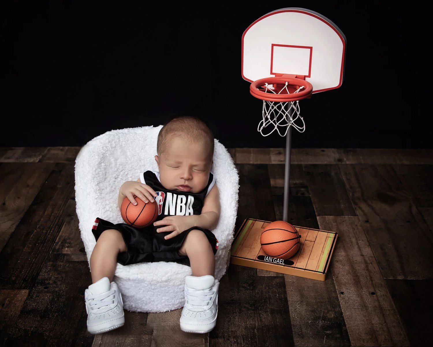 A sleeping baby in a black NBA basketball jersey and white sneakers, sitting in a white fuzzy chair, holding a small basketball. Next to the baby, there is a small basketball hoop with an orange backboard and net, and a basketball on the wooden floor